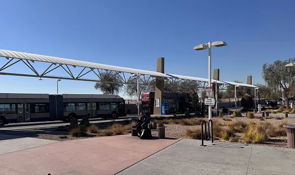 A sunny outdoor bus station with a long white canopy, parked buses, lamp posts, desert plants, and a person sitting on a bench. Clear blue sky and modern shelter structures are visible.