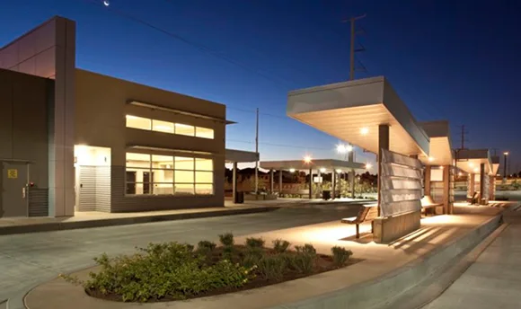A modern, well-lit bus transit station at dusk features covered waiting areas with benches, landscaped greenery, and a nearby building with large windows. Streetlights illuminate the quiet, empty platform.