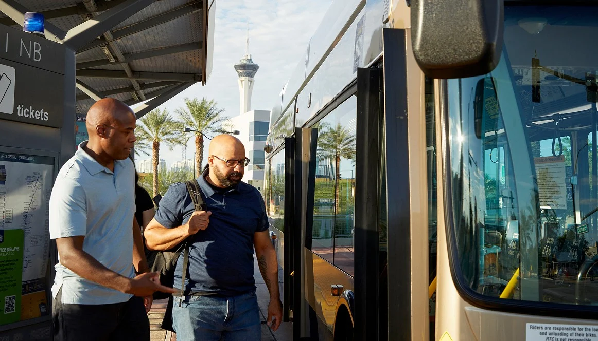 Two men stand at a bus stop, one gesturing while talking to the other, who wears a backpack. A bus is parked in front of them, and palm trees and a tall tower are visible in the background.