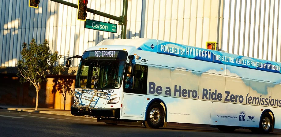 A blue and white RTC Transit bus promoting hydrogen power and zero emissions drives through a city street under a traffic light and a Carson street sign.