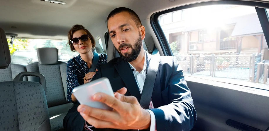 A man in a suit sits in the front seat of a car, holding a smartphone, while a woman wearing sunglasses sits in the back seat, looking forward. Sunlight streams through the windows.
