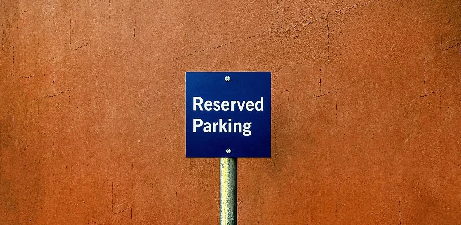 A blue sign with white text reading Reserved Parking is mounted on a metal pole in front of a textured orange wall.