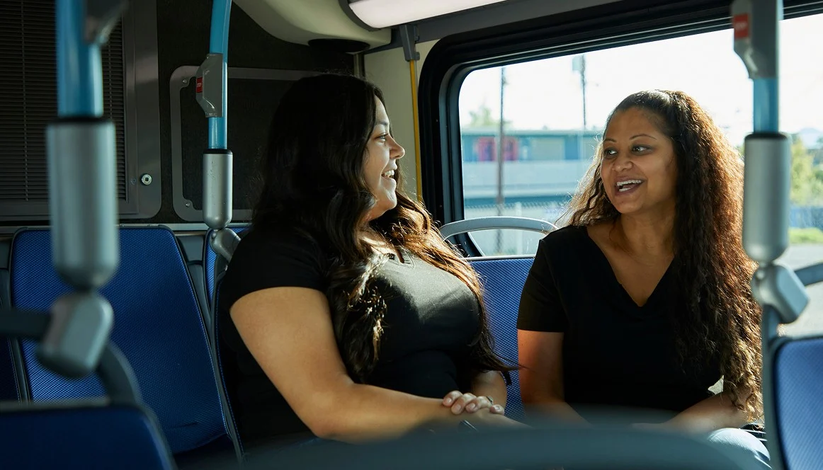 Two women sit on a bus, facing each other and smiling while having a conversation. The seats around them are empty, and sunlight shines through the window behind them.
