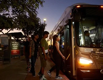 Four people are boarding a city bus at dusk. The sky is darkening, and the bus lights are on. Trees and a bus stop with a sign are visible in the background.
