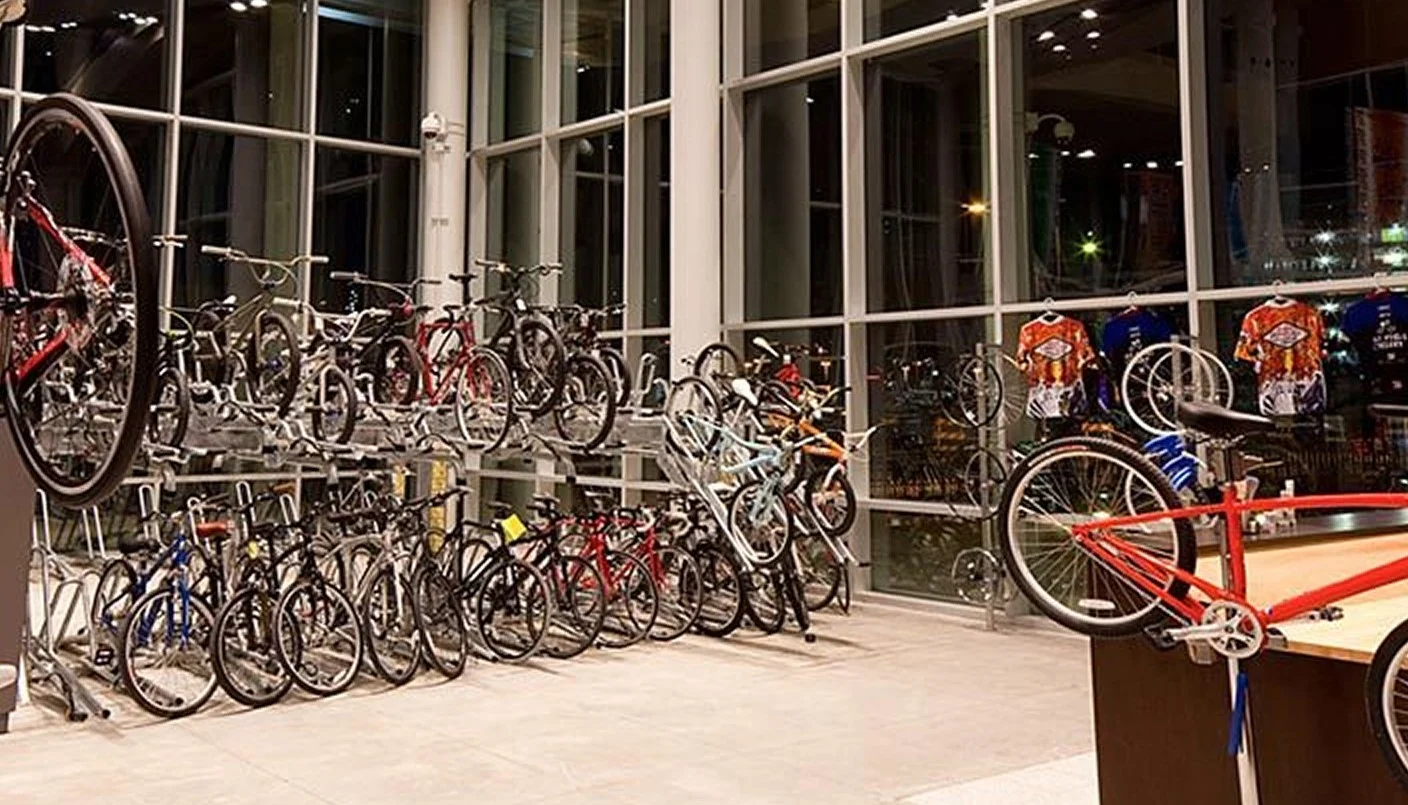 A modern bike shop interior with rows of bicycles on display racks, cycling jerseys hanging on the wall, and large windows showing night city lights outside.