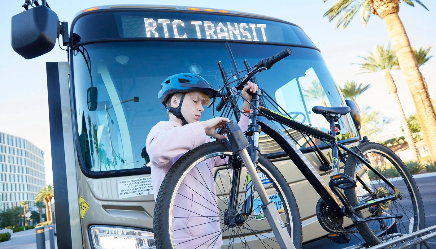 A person wearing a blue helmet loads a bicycle onto the front rack of an RTC Transit bus on a sunny day, with palm trees and modern buildings in the background.