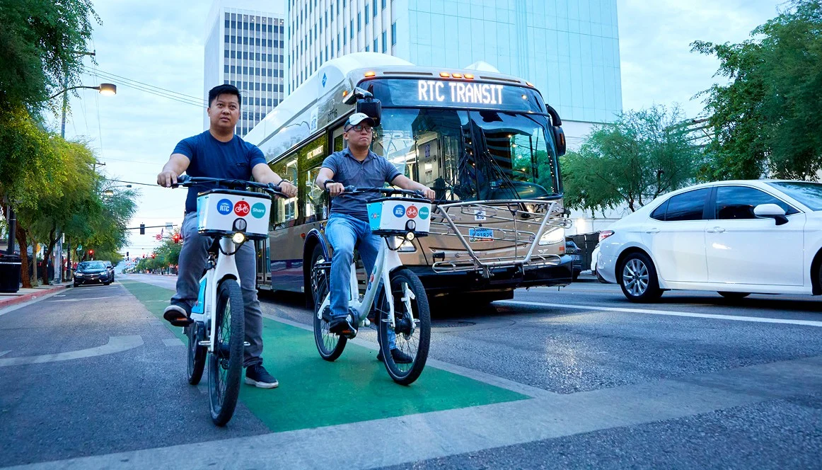 Two men ride rental bikes in a green bike lane on a city street, with a public transit bus and cars nearby. Tall buildings and trees line the background on a cloudy day.