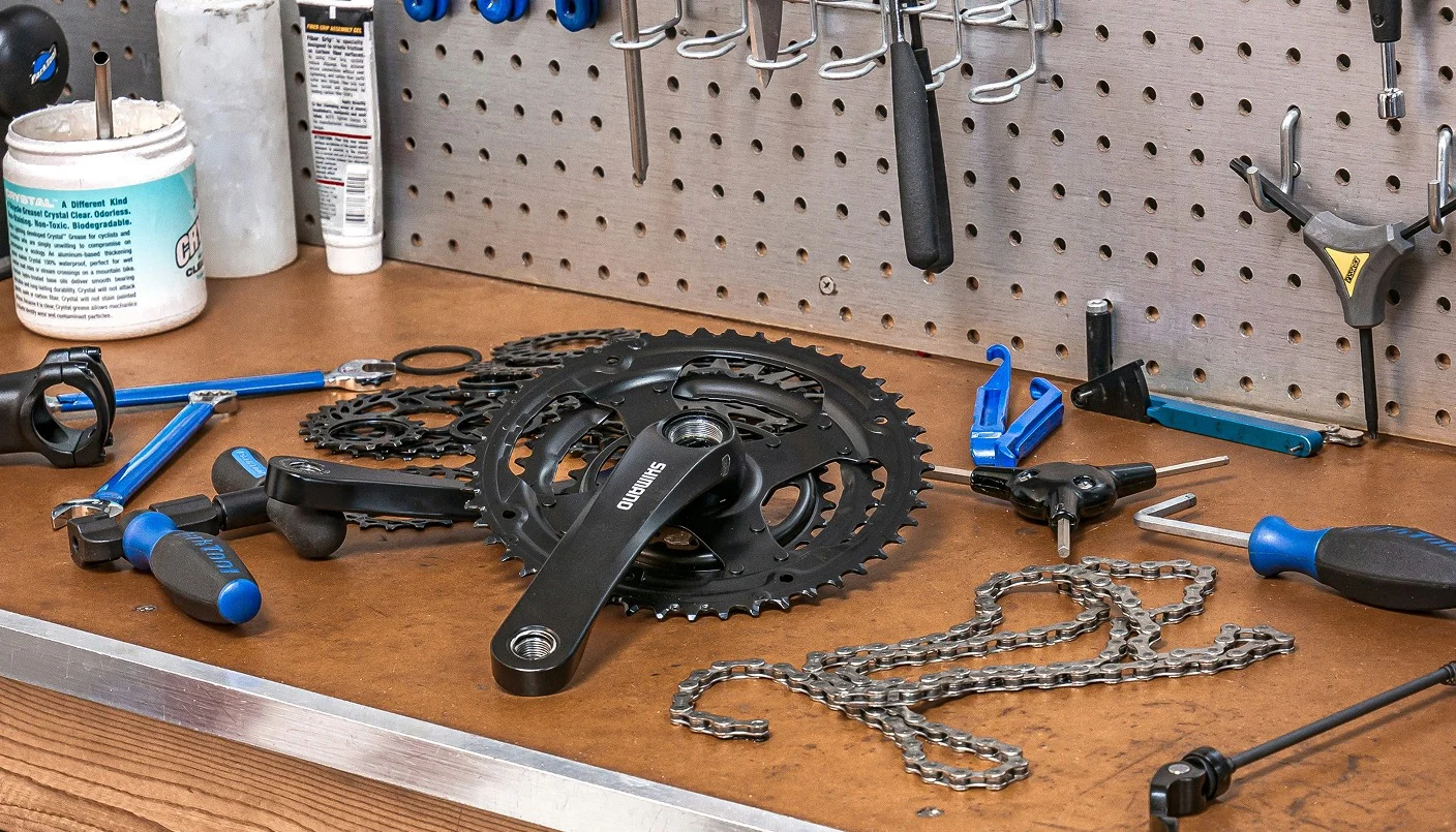 A workbench with bicycle repair tools, a bike chainring, a loose bike chain, gears, screwdrivers, and wrenches, with additional tools hanging on a pegboard in the background.