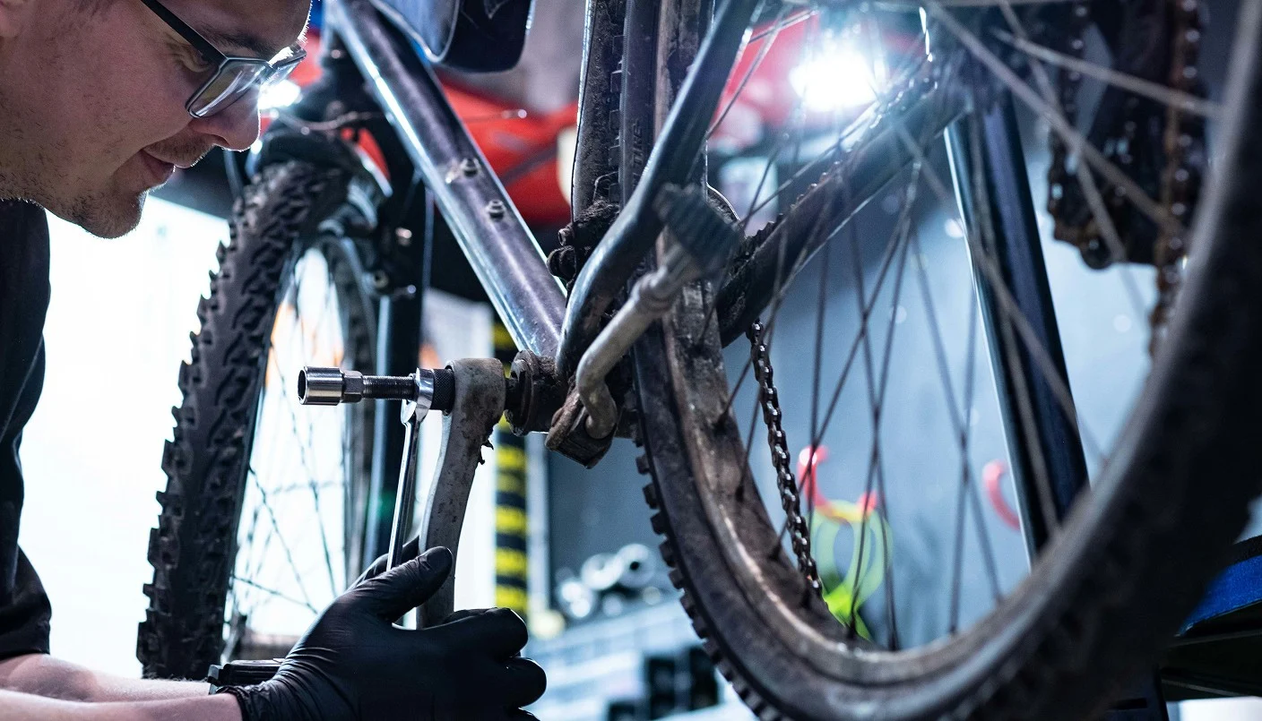 A person wearing glasses and black gloves repairs a mountain bike, focusing on the crank area, in a workshop with tools and bike accessories visible in the background.