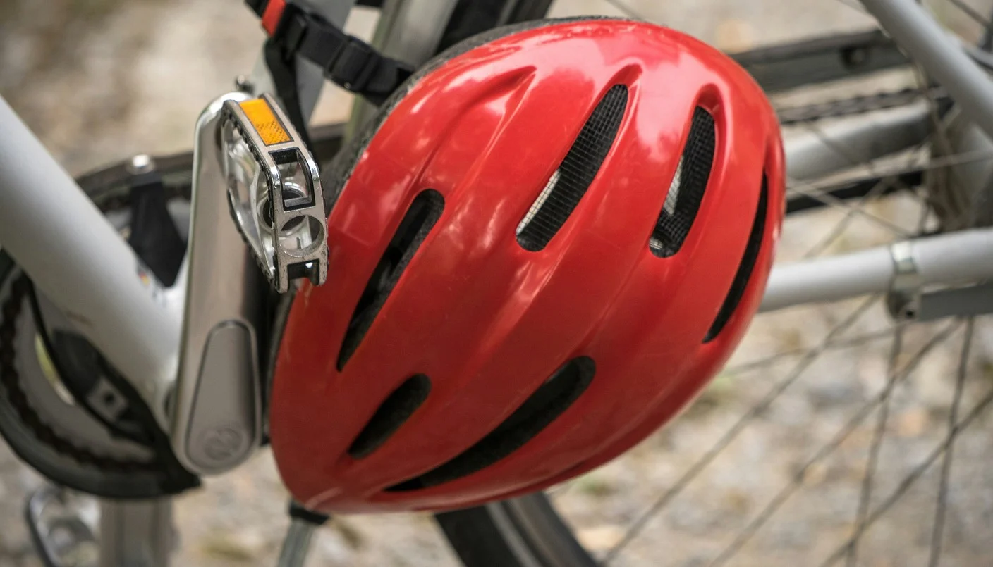A red bicycle helmet hangs from the frame of a silver bicycle, with part of the bike’s pedal, chain, and wheel visible in the background.