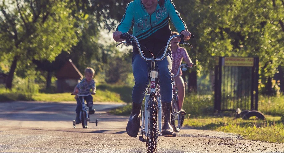 Three children ride bikes and a tricycle down a sunlit, tree-lined rural road. The child in front smiles while splashing through a puddle, with two other kids following closely behind.