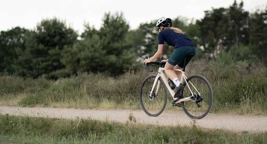A person wearing a helmet and cycling gear rides a bicycle on a gravel path through a grassy, wooded area. Trees and greenery are visible in the background under a cloudy sky.