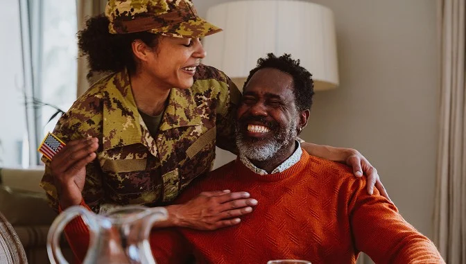 A smiling woman in military uniform embraces a cheerful man in an orange sweater, both seated indoors. The woman has her hand on the mans shoulder, and they appear to be sharing a joyful moment together.