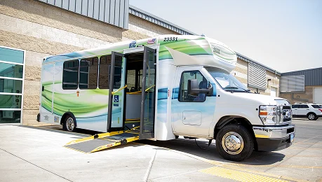 A white and green public transit bus parked outside a building with its door open, showing an extended wheelchair ramp for accessibility.