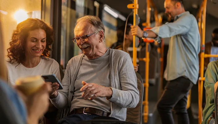 An older man and a woman smile while looking at a smartphone together on a bus. The man is laughing, and other passengers are visible in the background. Warm sunlight shines through the windows.