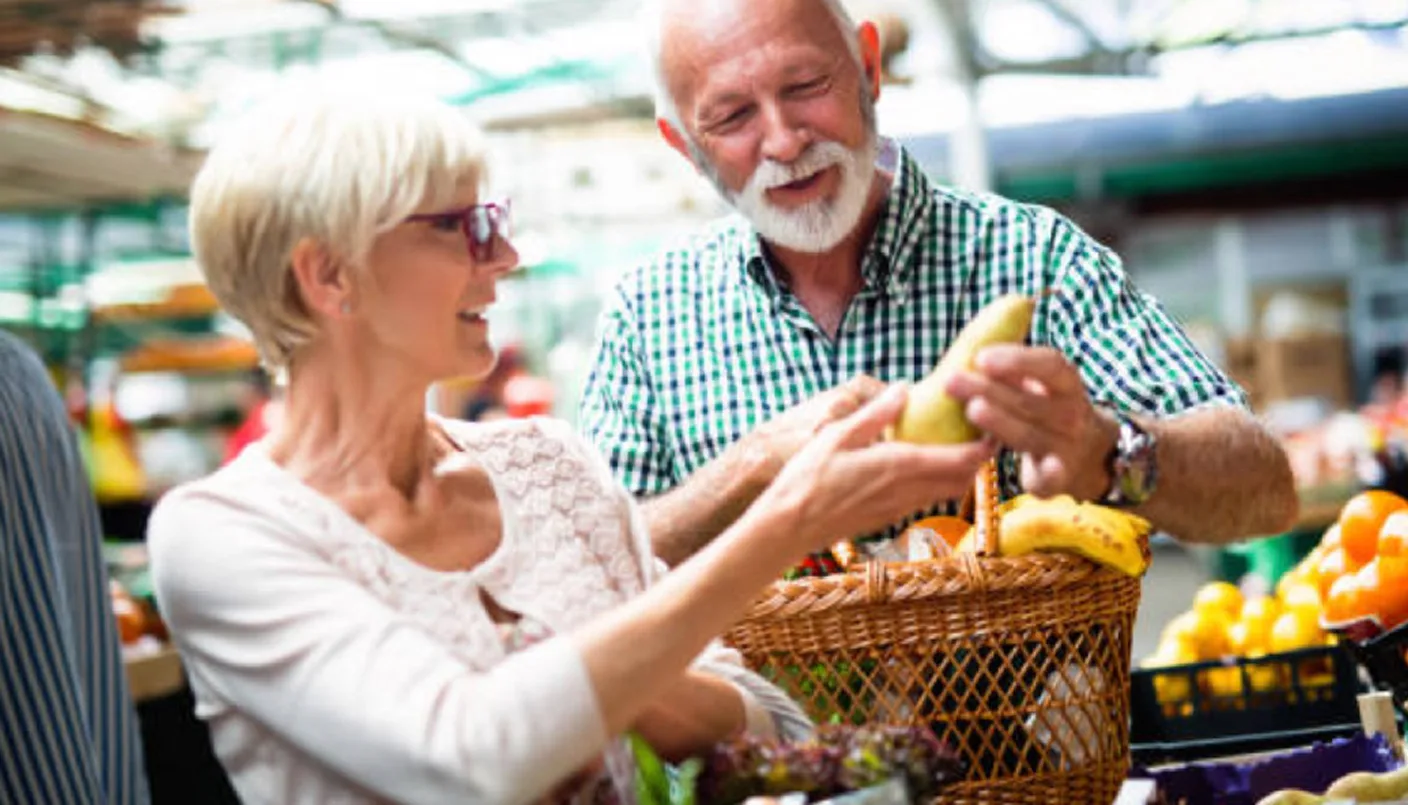 An older couple smiles while shopping for fresh produce at a market. The man holds a pear, and the woman, wearing glasses, looks at it. A shopping basket filled with fruits and vegetables is in her arm.