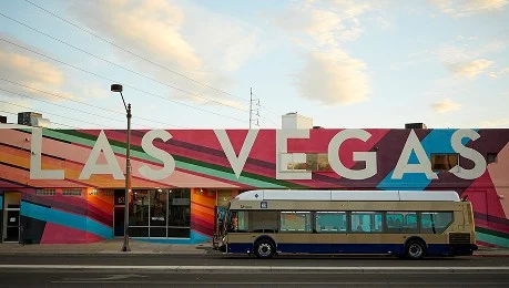 A colorful mural with large letters spelling LAS VEGAS covers a building wall, while a city bus is parked on the street in front of it under a partly cloudy sky.