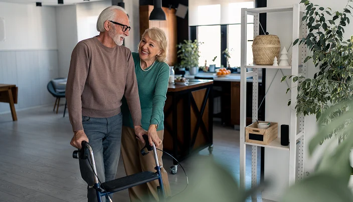 An elderly man using a walker smiles while his cheerful partner stands beside him, offering support in a bright, modern living space decorated with plants and shelves.