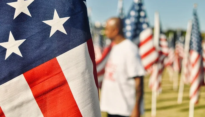 Close-up of an American flag in the foreground, with numerous other U.S. flags and a person standing in the blurred background, outdoors on a sunny day.