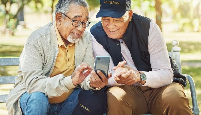 Two older men sitting on a bench outdoors, smiling and looking at a smartphone together. Both wear glasses and casual clothes; one wears a cap that says VETERAN. The scene is bright and friendly.
