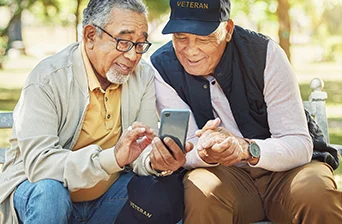 Two older men sit on a bench outdoors, smiling and looking at a smartphone together. Both wear glasses, and one is wearing a cap that says VETERAN. They appear to be enjoying each others company.