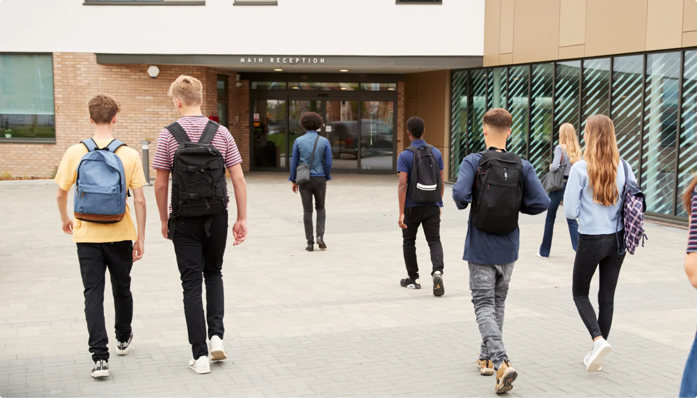A group of students with backpacks walk toward the entrance of a modern school building, heading for the main reception.