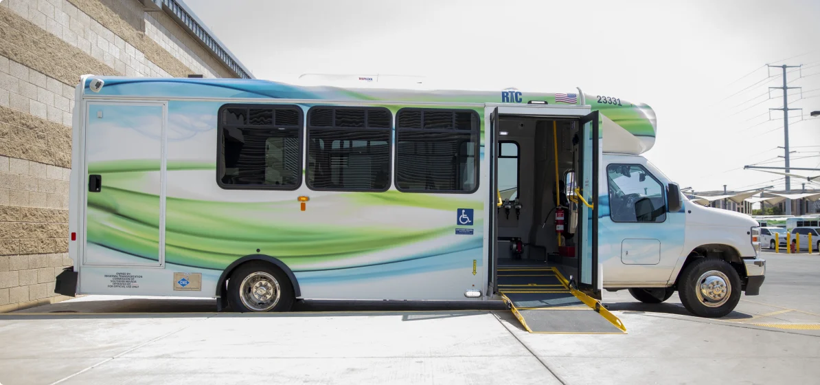 A white and green accessible transit bus parked with its door open and a yellow wheelchair ramp extended onto the sidewalk. The bus has large windows and is next to a beige brick building.