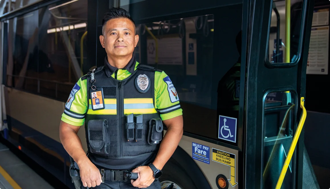 A transit security officer in a high-visibility uniform stands confidently at the entrance of a bus, which displays accessibility and fare signs near the door.