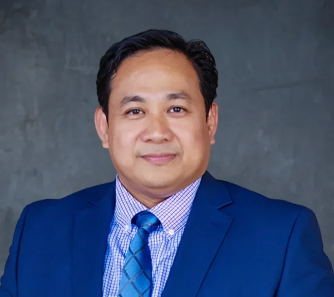 A man wearing a blue suit, patterned shirt, and blue tie stands in front of a gray textured background, smiling slightly at the camera.