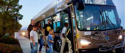 A group of people, including adults and children, are boarding a brown RTC Transit bus at a bus stop on a clear day. Trees and flowers line the sidewalk.