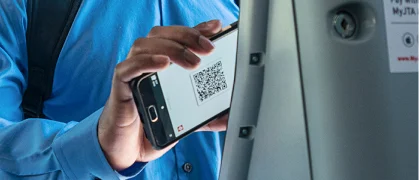 A person in a blue shirt holds a smartphone displaying a QR code near a scanner, possibly making a digital payment or validating a ticket.