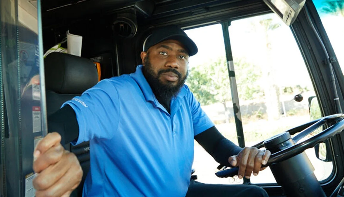 A man wearing a blue polo shirt and black cap sits in the drivers seat of a bus, holding the steering wheel with one hand and the bus door handle with the other, looking toward the camera.